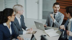 A team of four business professionals in a bright, modern office sitting around a conference table with laptops and notebooks, engaged in a collaborative discussion.